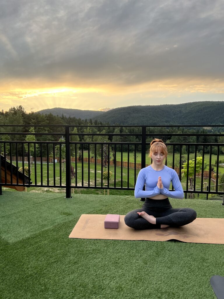 Kobieta praktykująca jogę na tarasie obiektu Serce Beskidu na tle zachodzącego słońca i panoramy gór. Zdjęcie ilustruje, jak sala warsztatowa w okolicach Krakowa może być rozszerzona o przestrzeń outdoor, idealną na sesje mindfulness, medytację czy treningi w otoczeniu natury.