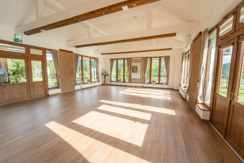 Widok na dużą, jasną salę warsztatową z drewnianą podłogą, belkami stropowymi i licznymi przeszkleniami wychodzącymi na ogród. Jest to profesjonalna sala na warsztaty dla kobiet w Krakowie i okolicach w ośrodku rekreacyjno-wypoczynkowym Serce Beskidu w Słopnicach.