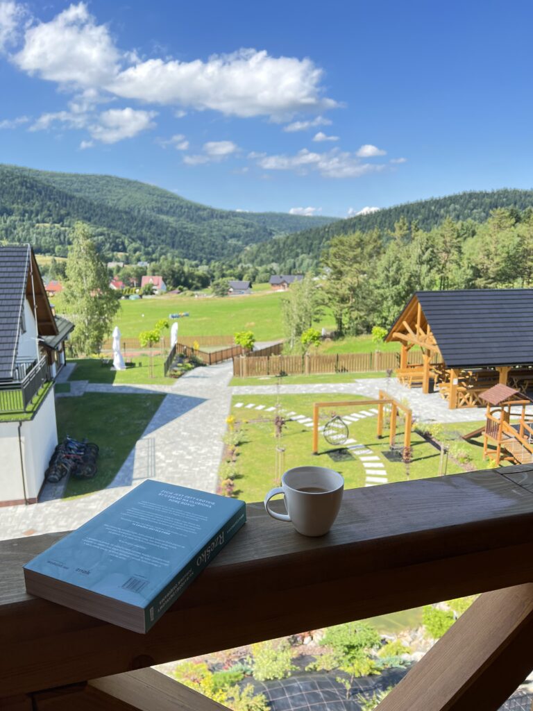 Widok z drewnianej balustrady na zadbany ogród ośrodka Serce Beskidu w Słopnicach w Małopolsce z alejkami, wiatami i placem zabaw, z majestatycznymi górami w tle. Na pierwszym planie leży książka oraz biały kubek kawy, tworząc idealny klimat pod kurs medytacji w górach i poranną kontemplację natury.