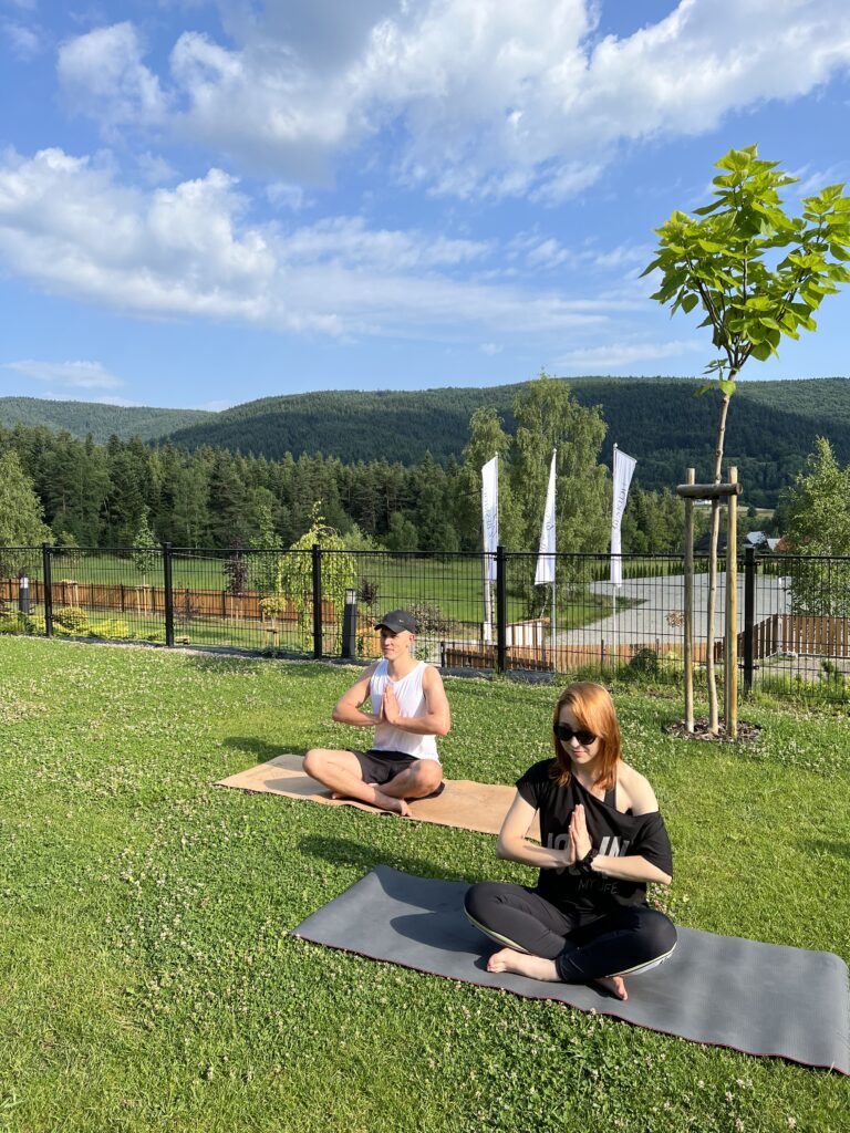 Kobieta i mężczyzna siedzą w pozycji lotosu na matach rozłożonych na zielonej trawie, praktykując uważność podczas warsztatu. W tle widać ogrodzenie obiektu Serce Beskidu, flagi reklamowe oraz zalesione pagórki, co idealnie uzupełnia weekendowy kurs medytacji w górach.