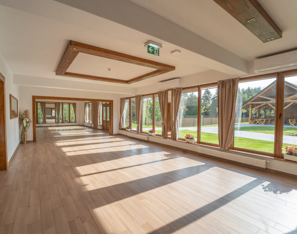 Widok na wnętrze jasnej, przestronnej sali treningowo-warsztatowej w Sercu Beskidu. Podłoga wyłożona jest jasnymi panelami, a sufit ozdabiają drewniane kasetony z wbudowanym oświetleniem LED i klimatyzatorami. Ogromne, sięgające od podłogi do sufitu okna tarasowe z beżowymi zasłonami wpuszczają mnóstwo naturalnego światła, oferując widok na zielony ogród. Ta nowoczesna przestrzeń idealnie obrazuje to, na co zwrócić uwagę, wybierając ośrodek na fit camp w Małopolsce – jakość sali ćwiczeń i warunki do pracy z ciałem.