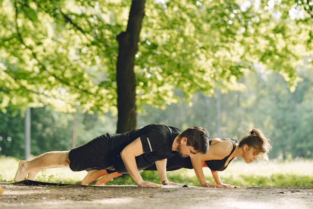 Kobieta i mężczyzna wykonują pompki na macie do ćwiczeń rozłożonej na ziemi w słonecznym lesie, co pokazuje, jak zorganizować fit camp blisko natury w otoczeniu ośrodka turystyczno-wypoczynkowego Serce Beskidu w Słopnicach.