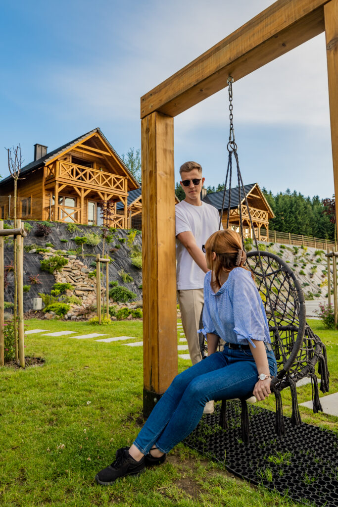 Nowoczesna, czarna huśtawka typu bocianie gniazdo zawieszona na solidnej, drewnianej ramie w ogrodzie Serca Beskidu. Siedzi na niej kobieta w błękitnej bluzce i jeansach, patrząca w stronę mężczyzny w białym t-shircie, który stoi obok, opierając się o belkę. W tle, na zadbanym, zielonym wzniesieniu, wznoszą się dwa luksusowe, całoroczne domki premium z bali, z tarasami i balkonami. Za nimi rozciąga się gęsty, ciemnozielony las iglasty pod jasnobłękitnym niebem. To sielskie ujęcie idealnie obrazuje najnowsze trendy w organizacji fit campu, który stawia na kameralne, nietypowe lokalizacje, komfortowe warunki noclegowe i bliskość natury, gwarantującą relaks i odcięcie od codziennych bodźców.