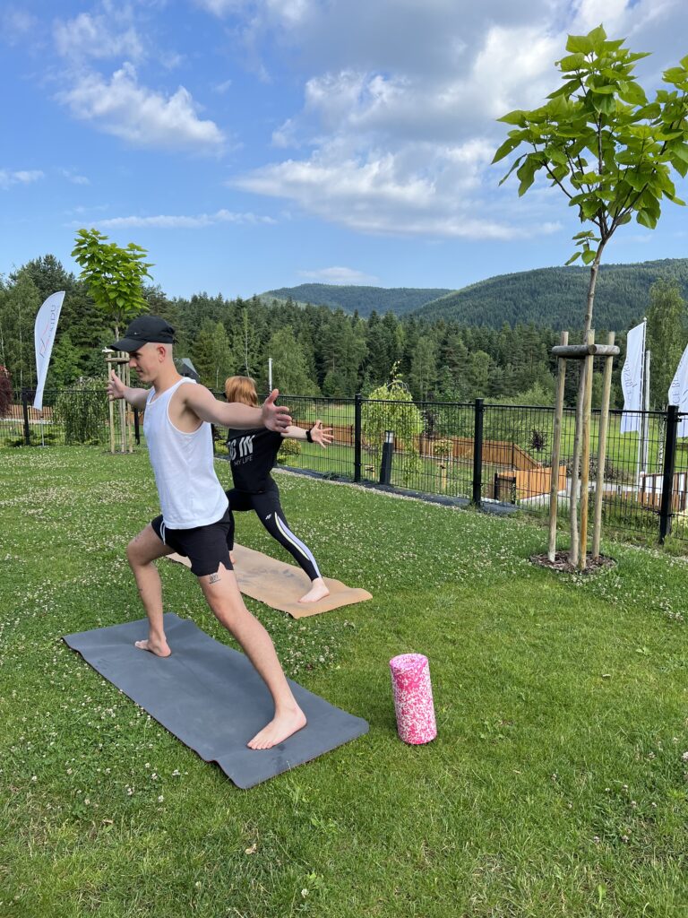 Mężczyzna i kobieta ćwiczą asany stojące na matach rozłożonych bezpośrednio na trawniku w ogrodzie Serca Beskidu. Joga w górach na świeżym powietrzu, z widokiem na zalesione szczyty, sprzyja lepszemu dotlenieniu organizmu i głębokiej relaksacji.