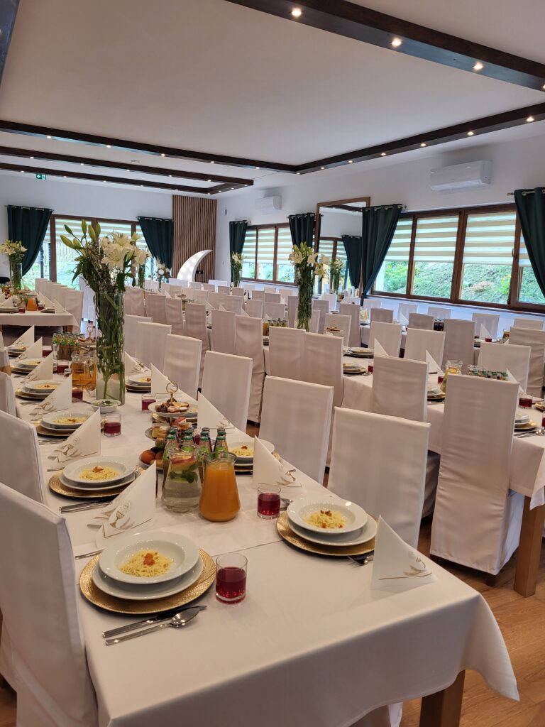 Przestronna sala w Serce Beskidu wypełniona elegancko nakrytymi stołami z białymi krzesłami, co stanowi idealną odpowiedź na dylemat: komunia w domu czy w lokalu. Zdjęcie pokazuje profesjonalne przygotowanie dużej uroczystości pod Krakowem, którego nie dałoby się zorganizować w domowych warunkach