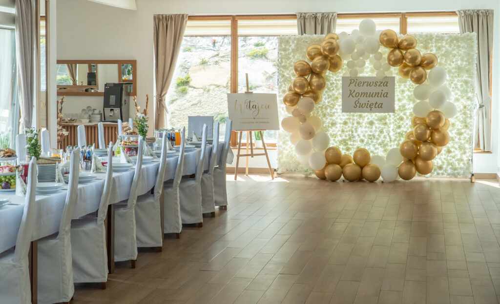 Jasne, nowoczesne wnętrze sali bankietowej w Serce Beskidu, jako alternatywa dla komunii w Krakowie na komunie w Krakowie Widoczne są długie stoły z białymi obrusami i pokrowcami na krzesła, a w tle duża ścianka dekoracyjna z białych kwiatów ozdobiona złotymi i białymi balonami z napisem „Pierwsza Komunia Święta”.
