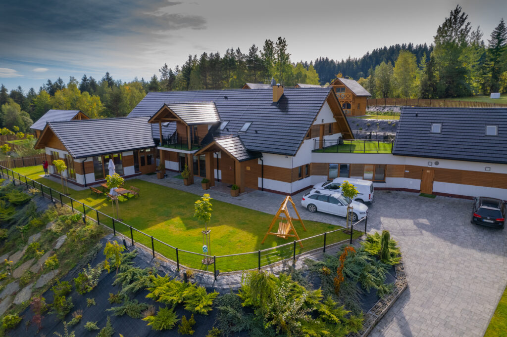 Widok z lotu ptaka na nowoczesny budynek Serca Beskidu w Słopnicach, otoczony zadbanym ogrodem z równo przystrzyżoną trawą. Szeroka przestrzeń zielona i tarasy to doskonałe warunki, aby uprawiać jogę na zewnątrz z widokiem na pasma górskie.