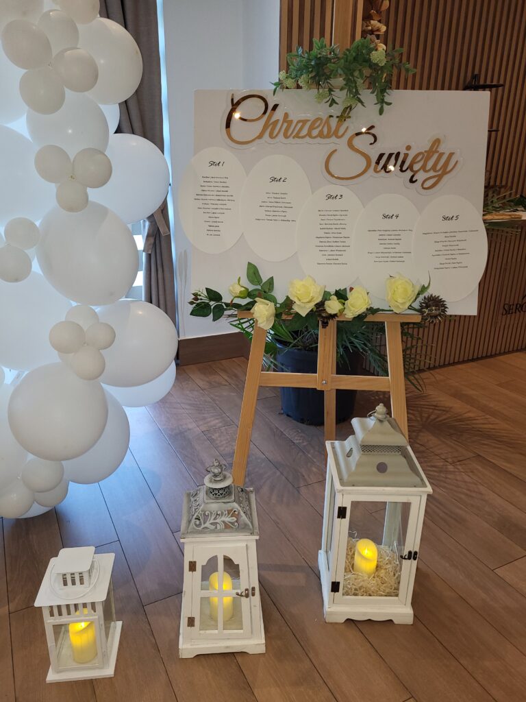 Stylowa tablica powitalna z imionami gości i dekoracyjnym napisem "Chrzest Święty" na sztaludze, otoczona balonami i lampionami, tworząca wyjątkowe dekoracje na chrzciny w restauracji Serce Beskidu.