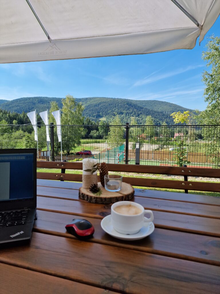 Stanowisko pracy zorganizowane na świeżym powietrzu na drewnianym tarasie w Sercu Beskidu. Na ciemnobrązowym stole z grubych desek stoi czarny laptop marki ThinkPad oraz bezprzewodowa, czarno-czerwona myszka komputerowa. Obok filiżanki z kawą na białym spodeczku leży dekoracyjny, drewniany krążek z białą świecą, szyszkami i zieloną gałązką. Nad stołem rozłożony jest duży, biały parasol ogrodowy chroniący przed słońcem. W tle za siatkowym ogrodzeniem widać zielone boisko, wysokie drzewa i majestatyczne, zalesione pasmo górskie, co ułatwia decyzję, gdzie w Polsce na workation warto się wybrać.