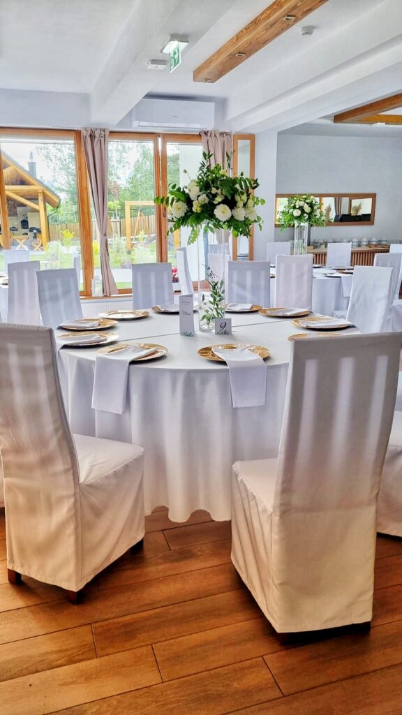 Elegancko nakryte okrągłe stoły z białymi obrusami i pokrowcami na krzesła w jasnej, nowoczesnej sali restauracji Serce Beskidu, przygotowane na przyjęcie z okazji chrztu świętego.