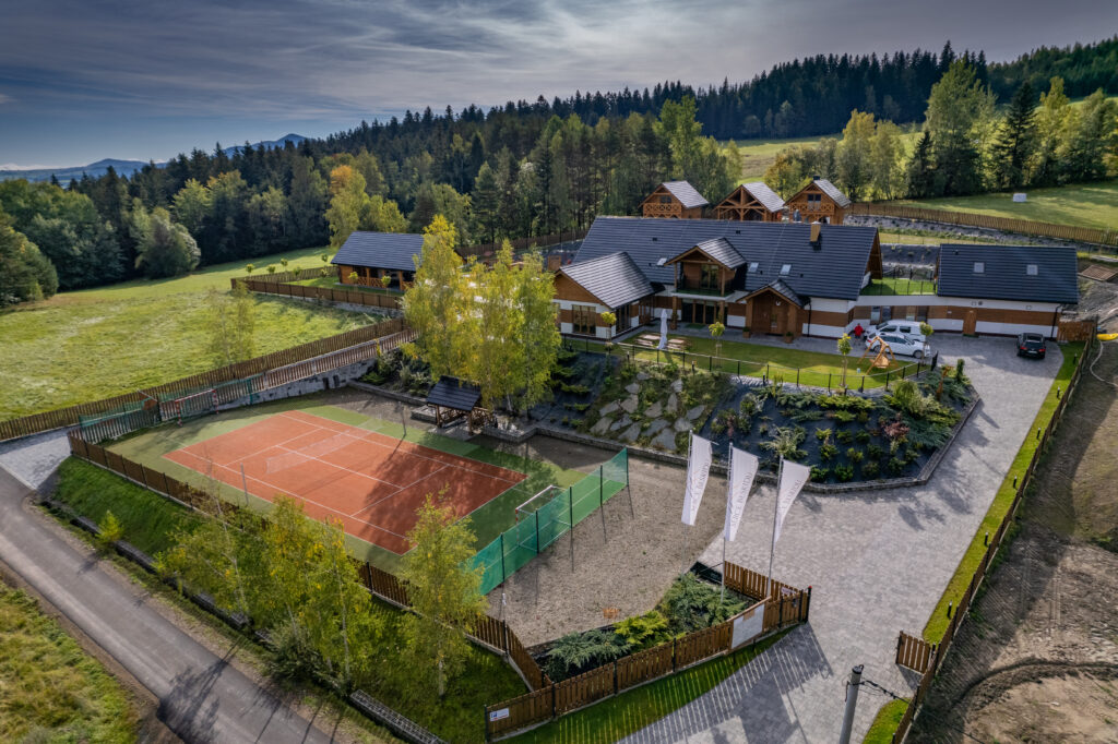 Widok z lotu ptaka na nowoczesny kompleks Serce Beskidu w Słopnicach, idealny na grupowe workation w górach. Na pierwszym planie widoczne jest profesjonalne, ogrodzone boisko sportowe o pomarańczowo-zielonej nawierzchni z bramkami, otoczone drewnianym płotem i młodymi brzozami. Obok boiska znajduje się wybrukowany parking z białymi flagami reklamowymi oraz zaparkowanymi samochodami osobowymi. W tle wznosi się duży, stylowy budynek główny z białymi ścianami i ciemnym, spadzistym dachem oraz mniejsze, drewniane domki całoroczne, otoczone gęstym lasem iglastym i zielonymi wzgórzami.