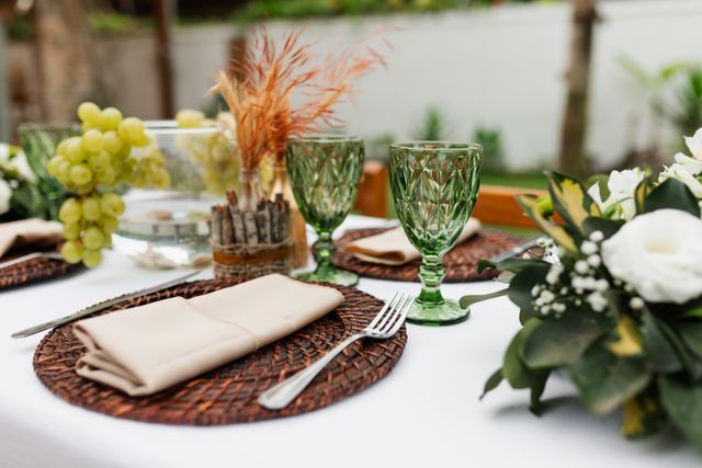 Zbliżenie na elegancko nakryty stół w obiekcie Serce Beskidu, przygotowany na wesele boho w górach Beskidach. Na białym obrusie widoczne są plecione podtalerze z ciemnego rattanu, na których spoczywają jasnobeżowe serwetki i srebrne sztućce. Uwagę przykuwają zdobione kieliszki do wina w głębokim, zielonym kolorze oraz centralna dekoracja składająca się z kiści świeżych winogron, suszonych traw w szklanych wazonach otulonych brzozową korą oraz bujnych kompozycji z białych kwiatów eukaliptusa.
