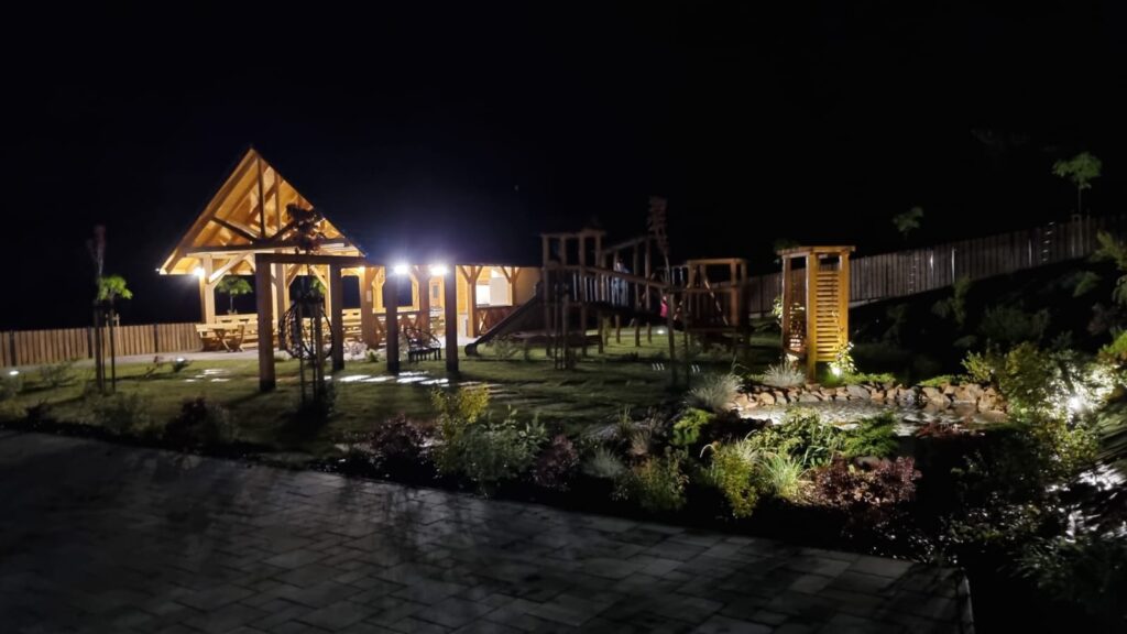 Nocny widok na zewnętrzny teren obiektu Serce Beskidu, gdzie oświetlona drewniana altana grillowa oraz plac zabaw tworzą bajkową atmosferę dla gości. Oświetlenie punktowe rozjaśnia ścieżki i roślinność, sprawiając, że ogród staje się romantycznym zakątkiem idealnym na wesele boho w Beskidach pod gwiazdami, z dala od zgiełku miasta.
