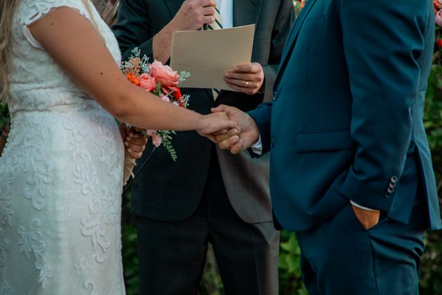 Zbliżenie na dłonie pary młodej splecione w czułym uścisku podczas ceremonii, którą zabezpiecza i organizuje Serce Beskidu. Po lewej stronie widoczna jest panna młoda od pasa w górę, ubrana w elegancką, białą suknię ślubną z koronkowym, florystycznym haftem i krótkimi rękawkami, która w drugiej dłoni trzyma bukiet ślubny złożony z łososiowych i różowych róż oraz drobnych, zielonych gałązek. Po prawej stronie stoi pan młody w dopasowanym, matowym garniturze w odcieniu ciemnego garnituru petrol blue lub głębokiego morskiego granatu, trzymający lewą dłoń w kieszeni spodni. W tle, pomiędzy małżonkami, widoczna jest sylwetka celebranta w ciemnym garniturze i krawacie w paski, który trzyma w dłoniach beżowy arkusz papieru z tekstem przysięgi i przemawia do zgromadzonych, a w tle majaczy naturalna, zielona roślinność.