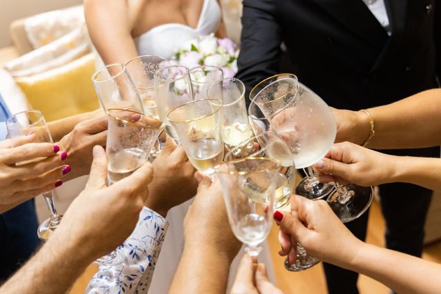 Zbliżenie na dłonie Pana Młodego, Panny Młodej oraz zebranych gości wznoszących wspólny toast eleganckimi kieliszkami z szampanem podczas przyjęcia w obiekcie Serce Beskidu. W tle dostrzec można fragment białej sukni ślubnej, delikatny bukiet jasnych róż oraz ciemny garnitur Pana Młodego. Ciepłe ujęcie idealnie oddaje bliską, swobodną i radosną atmosferę, jaką wyróżnia się kameralne wesele w górach organizowane w gronie najbliższych osób.