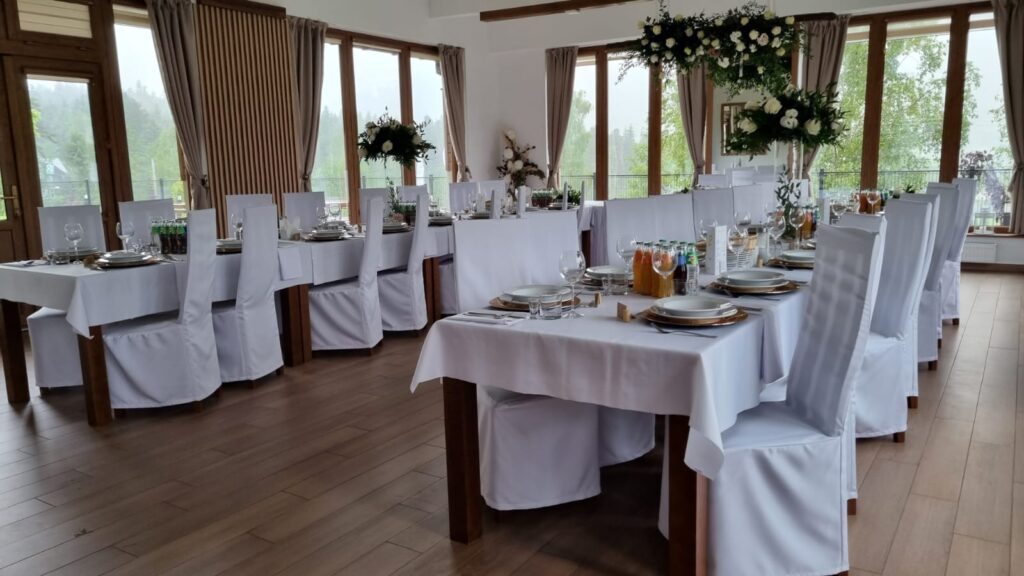 Jasna, elegancka sala weselna w obiekcie Serce Beskidu nakryta białymi obrusami i pokrowcami na krzesła. Na stołach ustawiono elegancką zastawę, wysokie kompozycje z białych kwiatów oraz napoje, a przez duże przeszklenia widać górski krajobraz. To idealne miejsce na kameralne wesele w górach dla 40 osób.