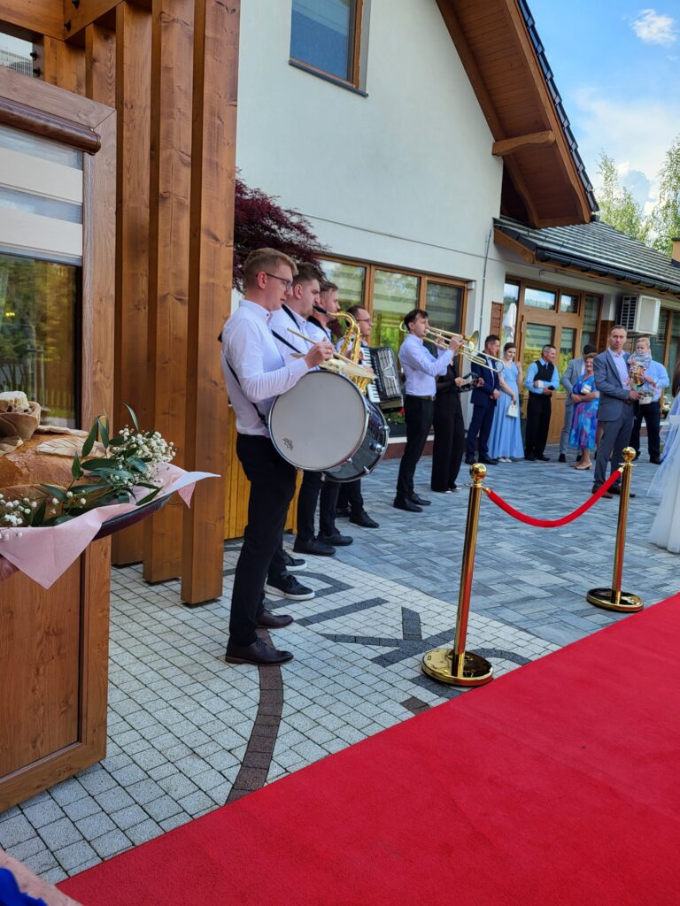 Pięcioosobowy zespół muzyczny w białych koszulach i ciemnych spodniach gra na instrumentach (bęben, saksofon, akordeon, trąbka) na zewnątrz, przed budynkiem domu weselnego Serce Beskidu. Muzycy stoją na jasnej kostce brukowej obok rozłożonego czerwonego dywanu ze złotymi słupkami odgradzającymi. W tle widoczni są elegancko ubrani goście weselni oczekujący na parę młodą, a po lewej stronie na drewnianym elemencie konstrukcyjnym wyeksponowany jest tradycyjny chleb weselny ozdobiony liśćmi i białymi kwiatami na różowej serwecie.
