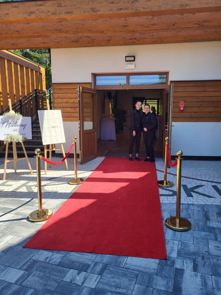 Dwie uśmiechnięte kobiety z obsługi w czarnych koszulach stoją w otwartych, drewnianych drzwiach wejściowych do nowoczesnego budynku w obiekcie Serce Beskidu. Przed wejściem rozłożony jest elegancki, długi czerwony dywan, odgrodzony złotymi słupkami z czerwonymi, sznurowymi barierami. Po lewej stronie na drewnianej sztaludze stoi biała tablica powitalna z napisem „Witamy”, udekorowana białymi kwiatami gipsówki. Po prawej stronie widoczna jest jasna ściana z elementami drewnianej szalówki oraz fragment napisu ułożonego z kostki brukowej na ziemi.