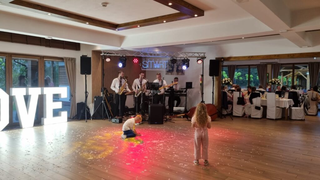 Czteroosobowy zespół muzyczny gra na instrumentach na podwyższeniu w nowoczesnej sali bankietowej obiektu Serce Beskidu, podświetlonej kolorowymi reflektorami z neonowym napisem „ZESPÓŁ SMART” w tle. Na drewnianym parkiecie rozsypane jest białe konfetti, na którym klęczy mały chłopiec w białej koszulce, a tyłem do obiektywu stoi dziewczynka w różowym kombinezonie z długimi, blond włosami. Po lewej stronie widoczny jest duży, podświetlany biały napis „LOVE”, a w głębi sali po prawej stronie znajdują się okrągłe stoły weselne przykryte białymi obrusami, przy których siedzą goście weselni.